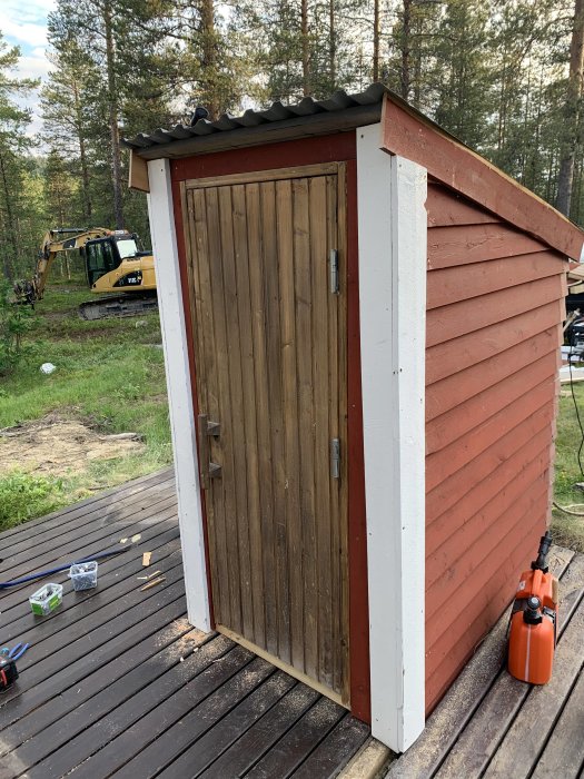 Träpumphus på trädäck med grävmaskin i bakgrunden, verktyg och sprejpump på marken.