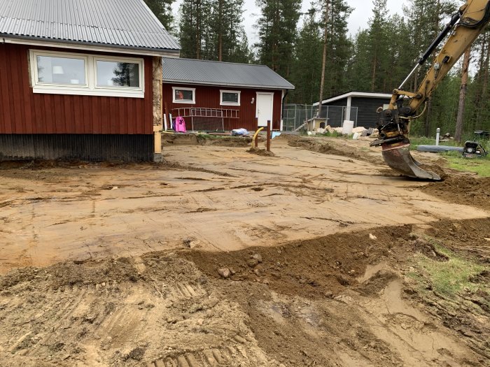 Grävmaskin som arbetar med att gräva nya vattenledningar vid en röd byggnad med ojämn, uppgrävd mark i förgrunden.