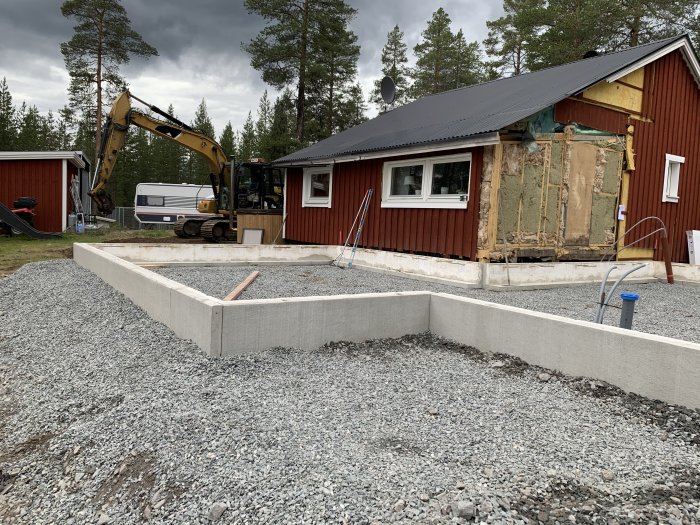 Grävmaskin bredvid hus med borttagen vägg, nygrundade kantbalkar och isolering.