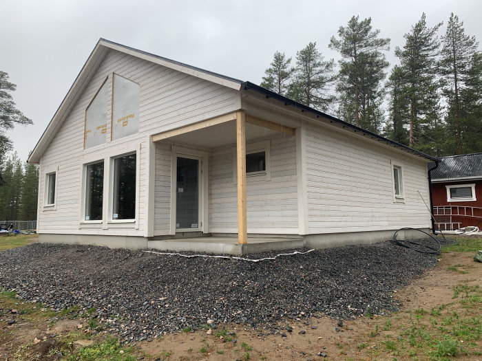 Nybyggd husutbyggnad med vit grundmålad panel, fönster och tak på plats, omgiven av grus och skog.