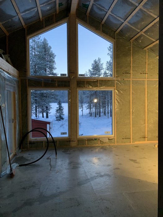 Inre utsikt av en nybyggd utbyggnad med isolering, fönster och dörrar installerade, sett mot en snöig skogsbakgrund.