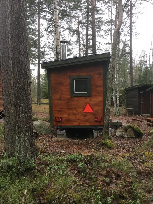 Mobil bastu i skogsmiljö med träpanel och skorsten, upphöjd på stödben med röda varningstrianglar på baksidan.