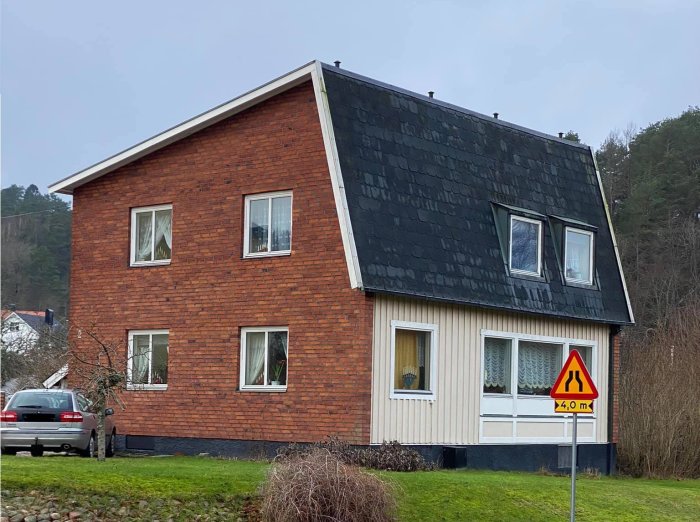 Tegelhus med ovanlig lutande takdesign i två våningar, trädgård, bil och vägmärke framför.