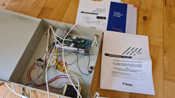 Nedmonterat larmsystem med centralenhet, kablar och instruktionsmanualer på trägolv.