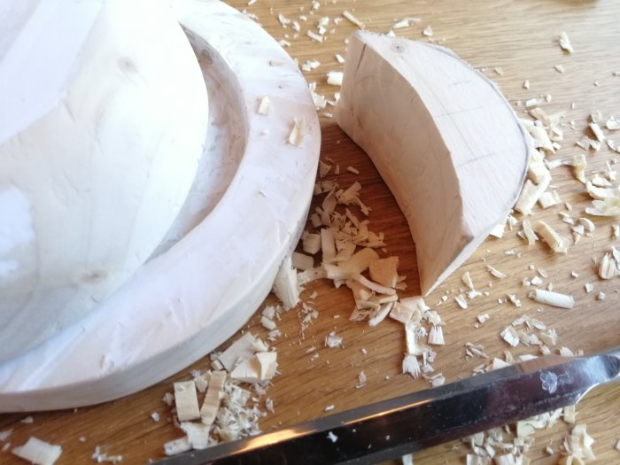 Ett separat snidat träföremåls huvud bredvid en bas med urfasning, omgivet av träspån och en snickarkniv.