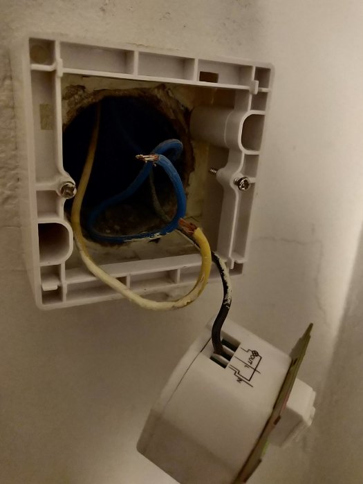 En öppen eluttagdosa i en vägg med en gammal strömbrytare hängande bredvid, och svarta, blåa och gula kablar synliga.