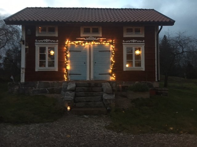 Röd trästuga med julbelysning runt dörren och fönster i skymningen.