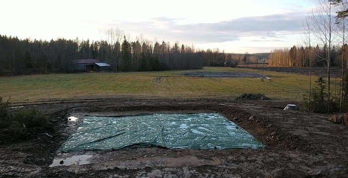 Nygrävd damm fylld med vatten bredvid en gräsbevuxen tomt och skog i bakgrunden.