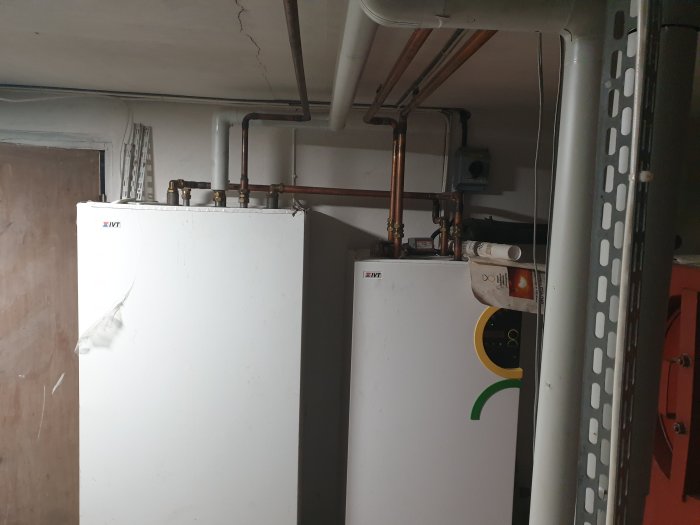 Två uppradade vita värmepumpstankar med kopparledningar och mätare synliga i ett tekniskt utrymme.