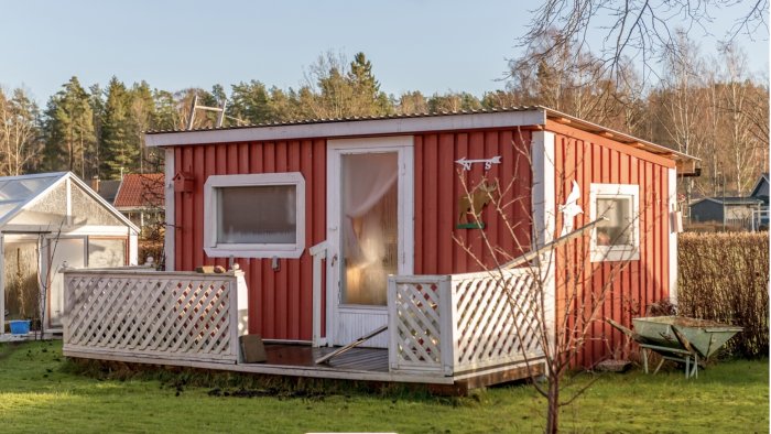 Litet rött hus på plintar med vit veranda och en lutning som indikerar sättning i marken.