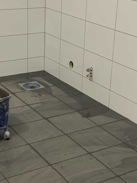Hörn av ett badrum med felplacerad golvbrunn i hörnet, vita kakelväggar och grå klinkergolv, samt rörutgångar på väggen.