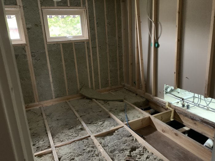 Interiör av ett pågående husbygge med exponerade träreglar, isolering i väggarna och ett öppet golv där rörinstallationer syns.