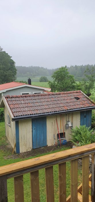 Utsikt från altan över ett gammalt uthus med slitet tak och blå dörrar omgivet av grönska.