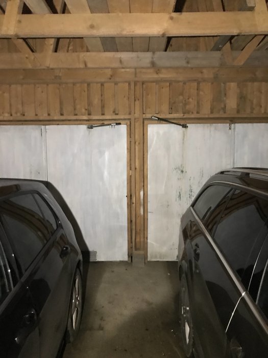 Två bilar parkerade i ett garage med träbjälkar och slitna vita dubbeldörrar i bakgrunden.