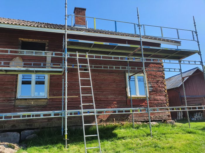 Ställning runt rödmålat trähus med synlig limträbalk och stenfot, under en klarblå himmel.