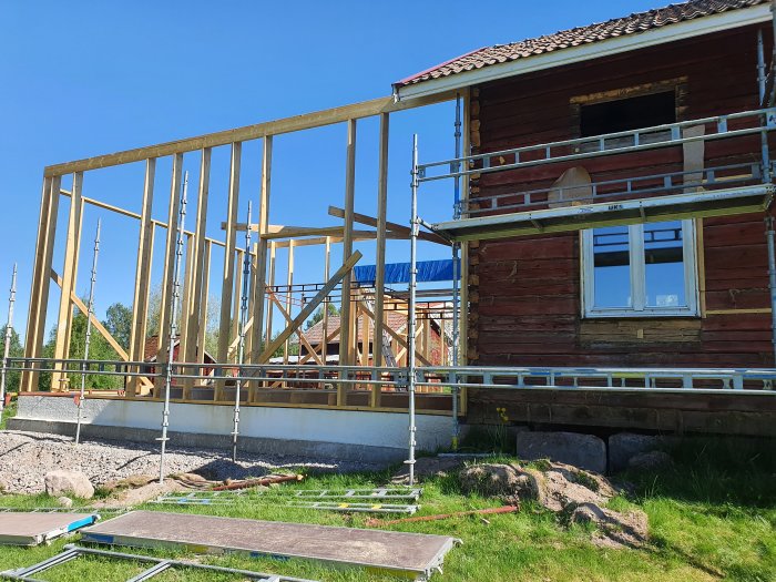 Byggställningar runt ett rött timmerhus med en limträbalk ovanför fönstret och en blå himmel i bakgrunden.