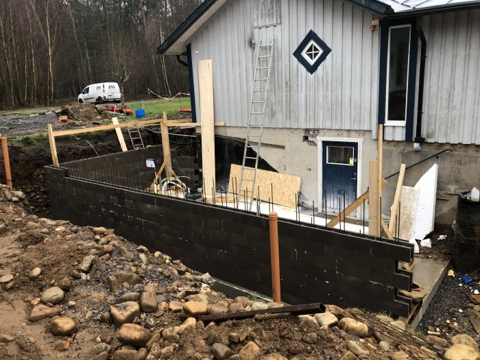 Byggarbetsplats med pågående arbete på en skalblocksmur, stödstag, och hus i bakgrunden.