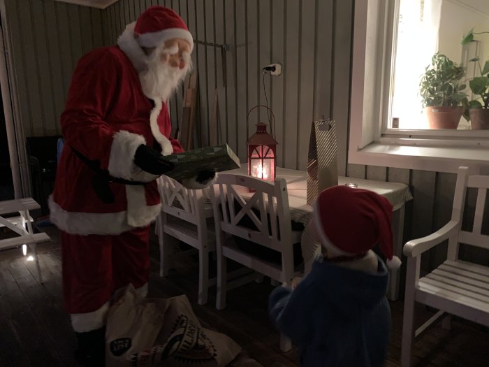 Person utklädd till tomte delar ut julklapp till barn på veranda i kvällsbelysning.