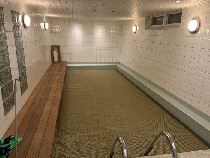 Inomhuspool med vita väggar och trätak, täckt av presenning, med synliga ventilationssystem.