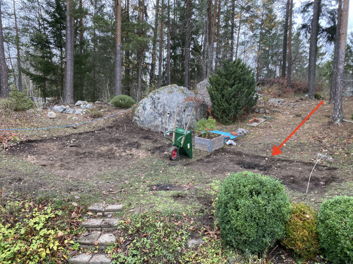 Trädgård med uthuggen mark för regnvattentank, trädgårdsredskap, och en röd pil som visar tankens planerade placering.