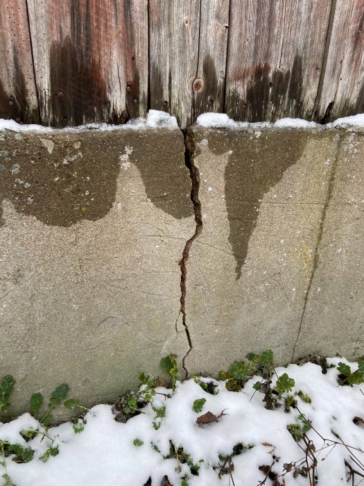 Sprucken grundmur med synlig spricka under trävägg, delvis täckt av snö.