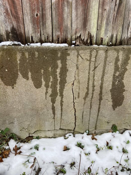 Sättningar i en ladugårds betonggrund med en spricka framför en vägg med slitet trä, snö intill.