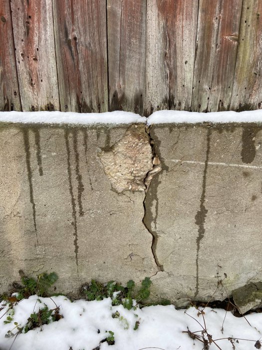 Sprucken gjuten betongplatta vid grund av ladugård med trävägg och snötäckt mark.