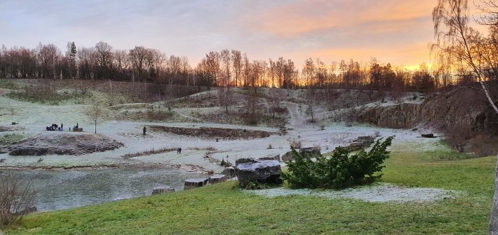 Vinterlandskap med frostdrabbat gräs vid soluppgång, personer samlade på en liten ö i en frusen damm.