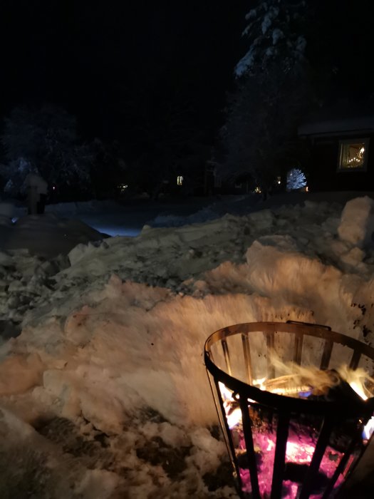 Eldkorg med brinnande eld ute i snön framför ett hus med upplysta fönster på kvällen.