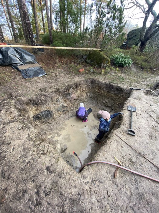 Två personer gräver i en lerig grop utomhus med spade och hink, omgivna av träd och verktyg.