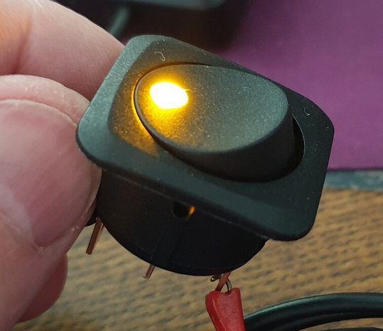 Aktiverad strömbrytare hållen i en hand, med lysande gul lysdiod och synliga terminaler.