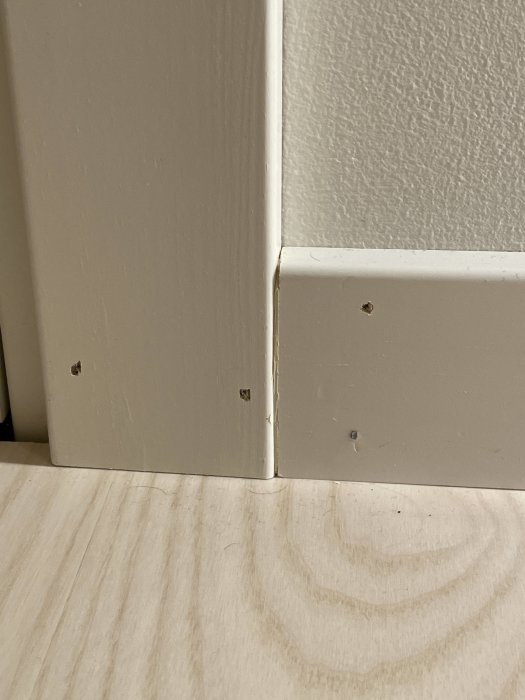 Vit fönsterlist och golvlist med spikhål, monterade mot en vägg och ett trägolv.