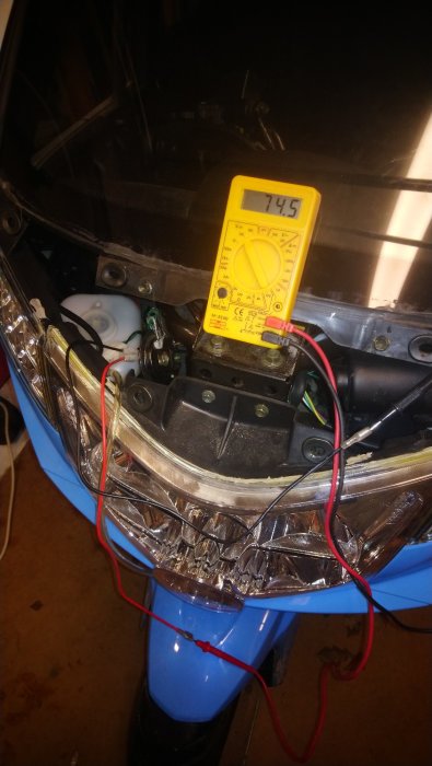 Multimeter som visar 74,5 volt ansluten till en moped med röd och svart kabel.