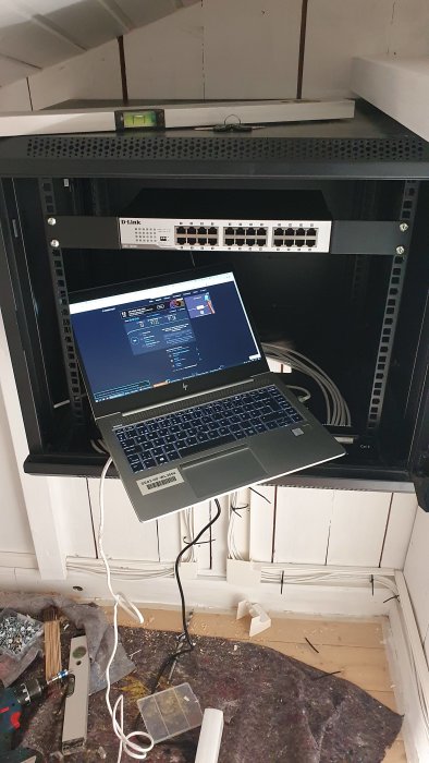 En öppen nätverksskåp med switchar och kabelrullar, ovanpå en HP laptop på en arbetsbänk under renovering med synliga kablar och verktyg.