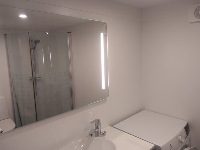 Nyinstallerad duschvägg i ett nyligen renoverat badrum med synlig tvättmaskin och upplyst spegel.