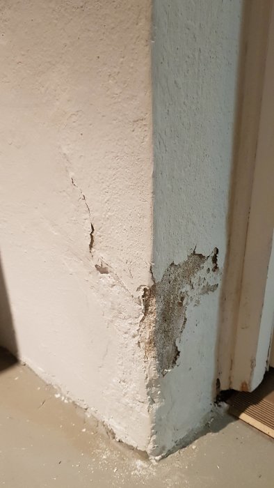 Skadad innervägg i källare med avflagad färg och sprickbildning vid hörnet och på den vita väggytan.