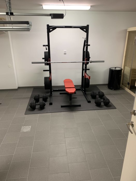 Hemgym med vikter, träningsbänk och squat rack på granitkeramikgolv, samt en synlig bastudörr.