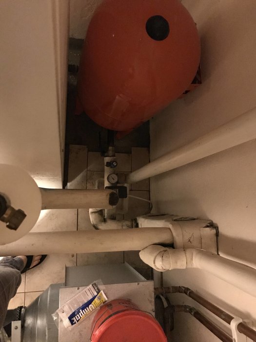 Rördetaljer och röd expansionskärl i pannrum med synlig isolering och underhållsprodukter på golvet.