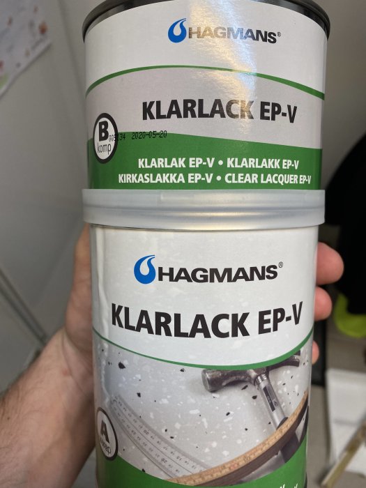 Hand håller en burk med Hagmans Klarlack EP-V, en epoxibaserad klarlack mot en oskarp bakgrund.