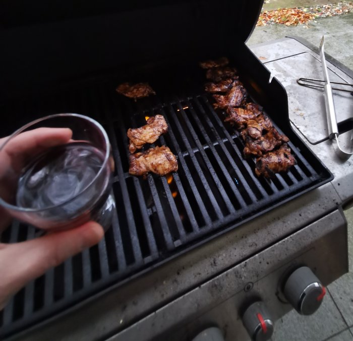 Grillat kött på en gasolgrill och en hand som håller ett glas vin.