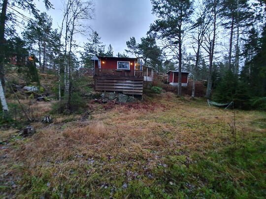 Stuga på höjd i skogsmiljö med veranda och skjul, gräsbevuxen mark i förgrunden.