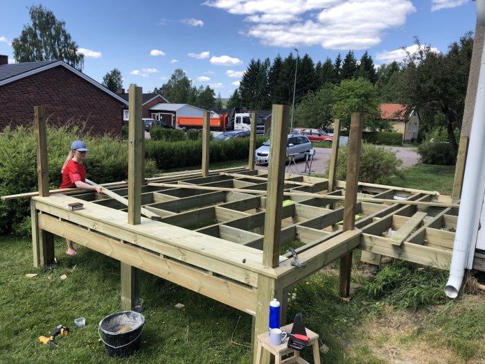 Nybyggd träkonstruktion av en altan med stolpar och bärlinor, och en person som arbetar på den.