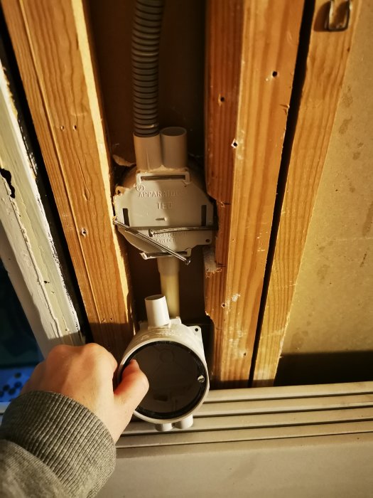 Hand håller en öppen apparatdosa vid en vägg med synliga elledningar och tomma rörinstallationer.