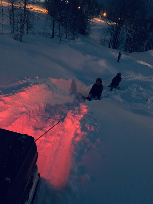 Två personer sitter i en snöig slänt vid skymning, dragna av ett fordon med rödbelysning underifrån.