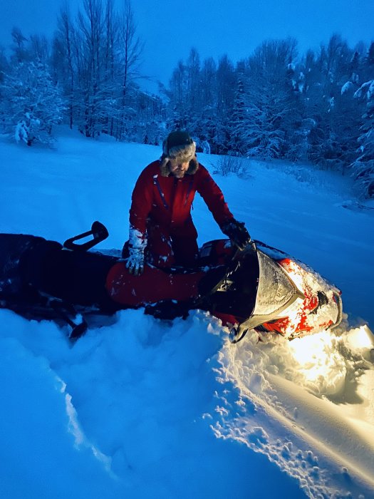 Person i röd vinterklädsel bredvid en snöskoter i snötäckt landskap under skymningen.