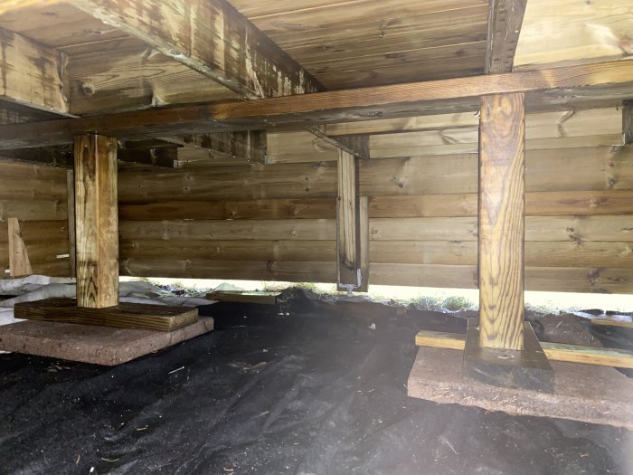 Stomme och stödben under en träkonstruktion med svart markduk och stenplattor som grund.