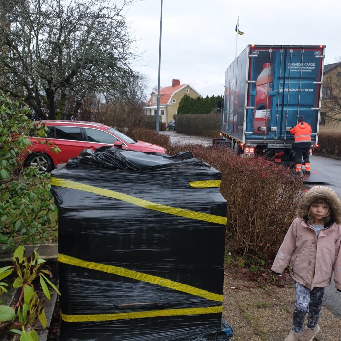 En pall med badrumsmöbler täckt av svart plast framför hus, lastbil och förare i bakgrunden.