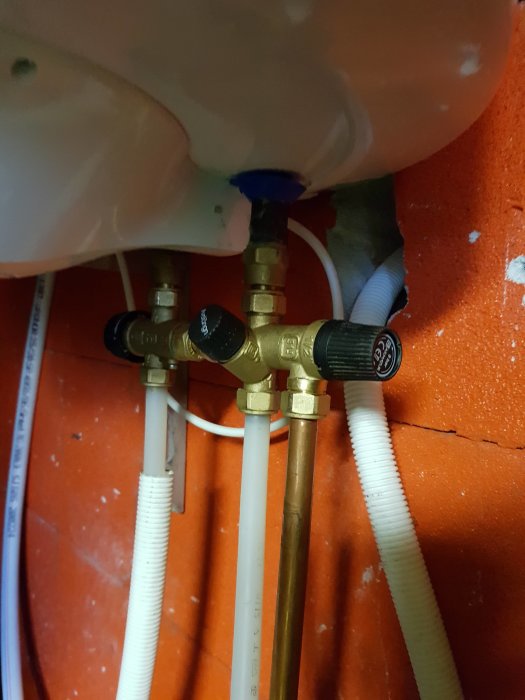 Röranslutningar och ventiler under en vit varmvattenberedare monterad på en röd vägg.