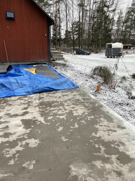 Nyjuten betongplatta täckt med en blå presenning vid kant, snö på marken, röd byggnad och hästtransport i bakgrunden.