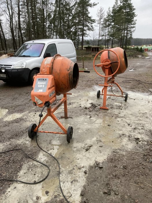 Två cementblandare på en lerig byggplats med skåpbil i bakgrunden.